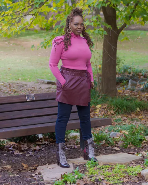 Antoinique standing by a park bench in a pink turtleneck, burgundy faux-leather skirt, and houndstooth ankle boots.