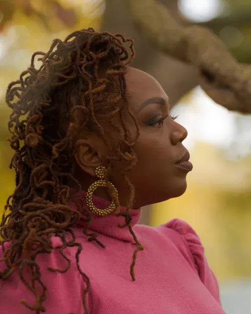 Antoinique in profile, pink turtleneck and gold hoop earring, looking out toward golden autumn leaves.
