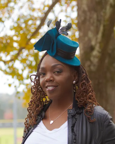 Antoinique smiling at the camera in a teal feathered fascinator hat and black leather jacket over a white tee, standing by a tree in autumn.
