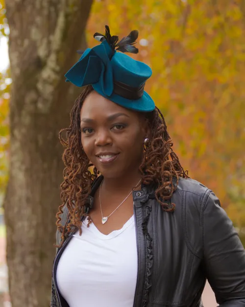 Antoinique smiling in the teal feathered fascinator hat and black leather jacket, framed by a tree trunk and autumn foliage.
