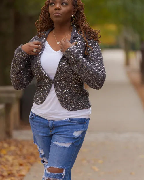 Antoinique walking along a tree-lined path in a sequined blazer, white tee, and ripped jeans, adjusting her lapels.