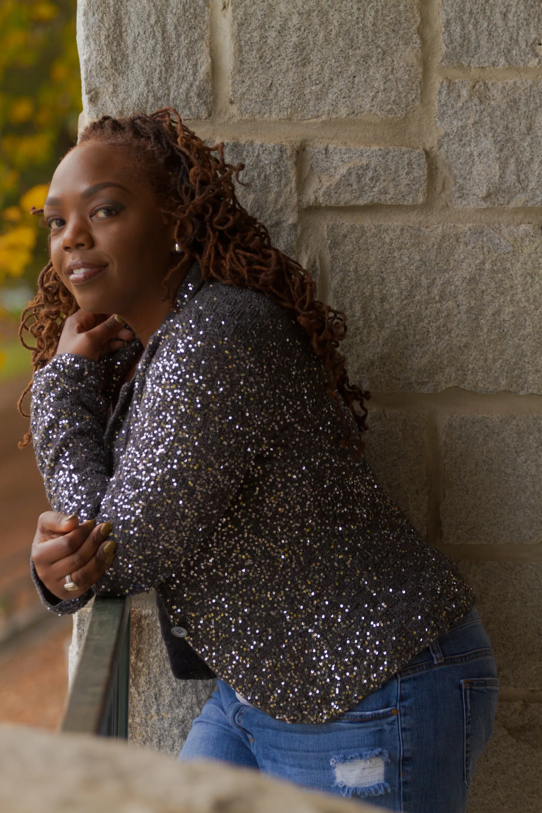Antoinique in a sequined charcoal blazer, leaning against a stone pillar with autumn leaves in the background.