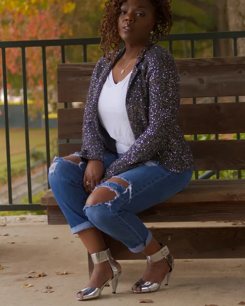 Antoinique seated on a wooden bench in a sequined blazer and cuffed ripped jeans, silver ankle-strap heels, autumn foliage behind a railing.