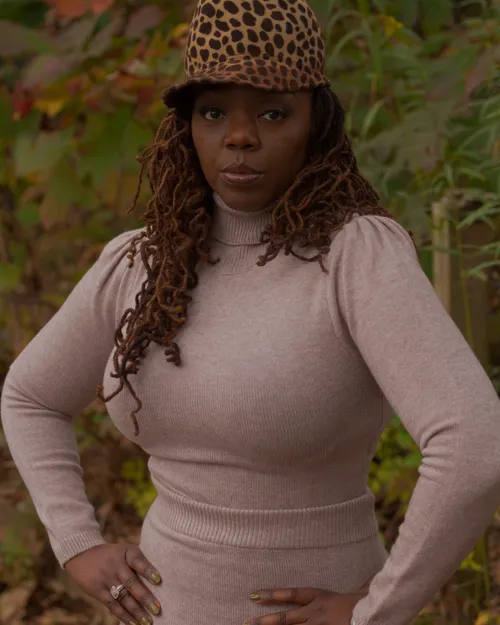 Antoinique standing with hands on hips in a leopard-print hat and blush turtleneck-and-skirt set, green foliage behind her.